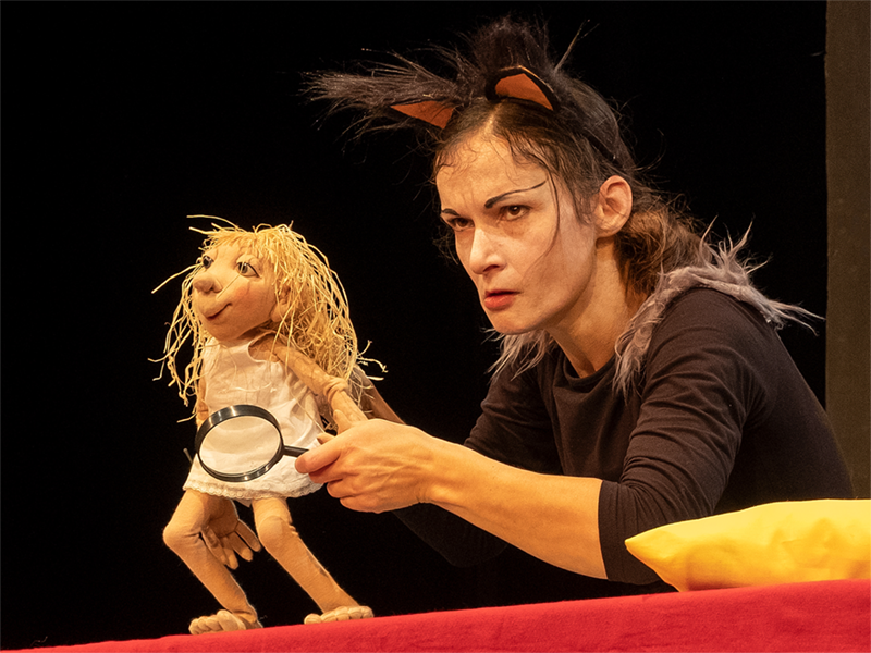 Puppenspielerin hält eine Lupe vor eine Strohfigur im weißen Kleid – Szene aus „Die Zeitdiebe“