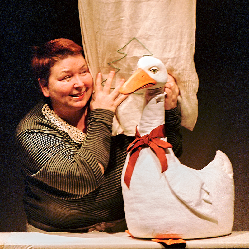 Puppenspielerin neben einer großen weißen Gans mit rotem Band – Szene aus „Die Weihnachtsgans Auguste“