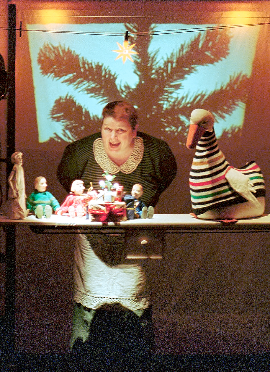 Spielerin hinter einer Bühne mit kleinen Figuren und einer gestreiften Gans, im Hintergrund ein projizierter Tannenbaum – Szene aus „Die Weihnachtsgans Auguste“
